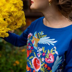 Puede incluir: Una camisa azul de manga larga con flores bordadas de colores y un dise&ntilde;o de p&aacute;jaro.