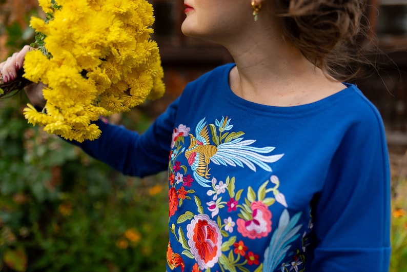Puede incluir: Una camisa azul de manga larga con flores bordadas de colores y un dise&ntilde;o de p&aacute;jaro.