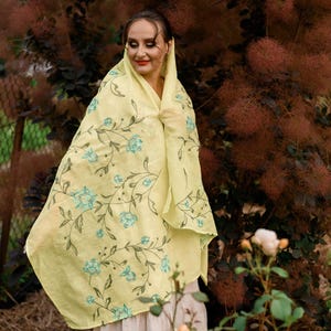 May include: A woman wearing a yellow floral scarf with a light blue floral pattern. The scarf is draped over her head and shoulders. She is standing in front of a bush with brown leaves.