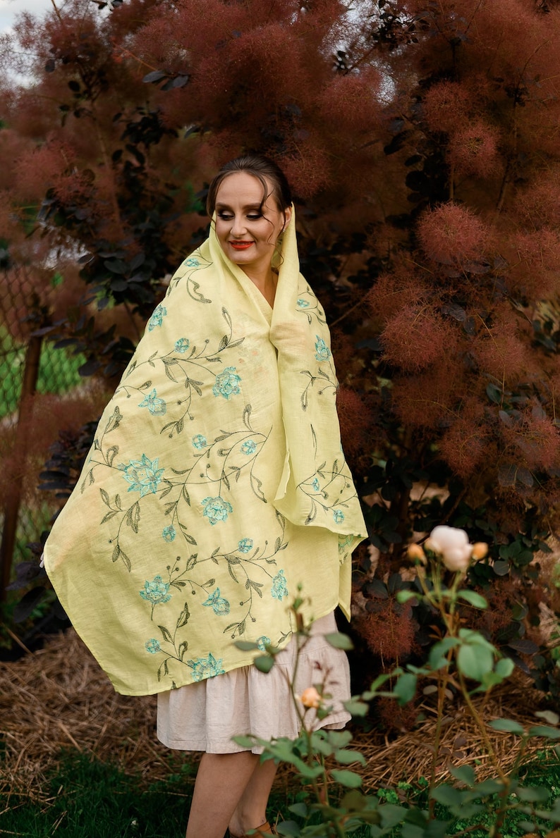 May include: A woman wearing a yellow floral scarf with a light blue floral pattern. The scarf is draped over her head and shoulders. She is standing in front of a bush with brown leaves.