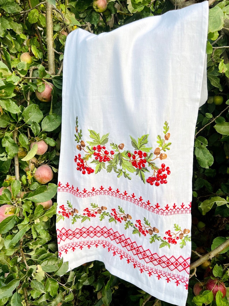 K&ouml;nnte beinhalten: Wei&szlig;es Leinenhandtuch mit gestickten roten Beeren, gr&uuml;nen Bl&auml;ttern und braunen Tannenzapfen. Der untere Rand hat ein rotes geometrisches Muster. Das Handtuch h&auml;ngt an einem Ast mit &Auml;pfeln.