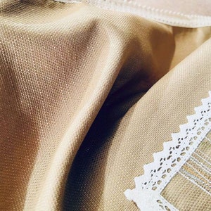 May include: Close-up of a beige fabric with a textured weave. White lace trim is visible along the edges, adding a delicate detail. The fabric is folded, creating shadows and highlighting the texture and color variations.
