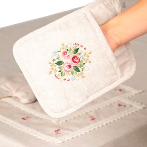 May include: Beige oven mitt with an embroidered floral design featuring pink roses, green leaves, and blue accents. The mitt is held over a matching tablecloth with embroidered flowers and lace trim, creating a cohesive kitchen textile set.