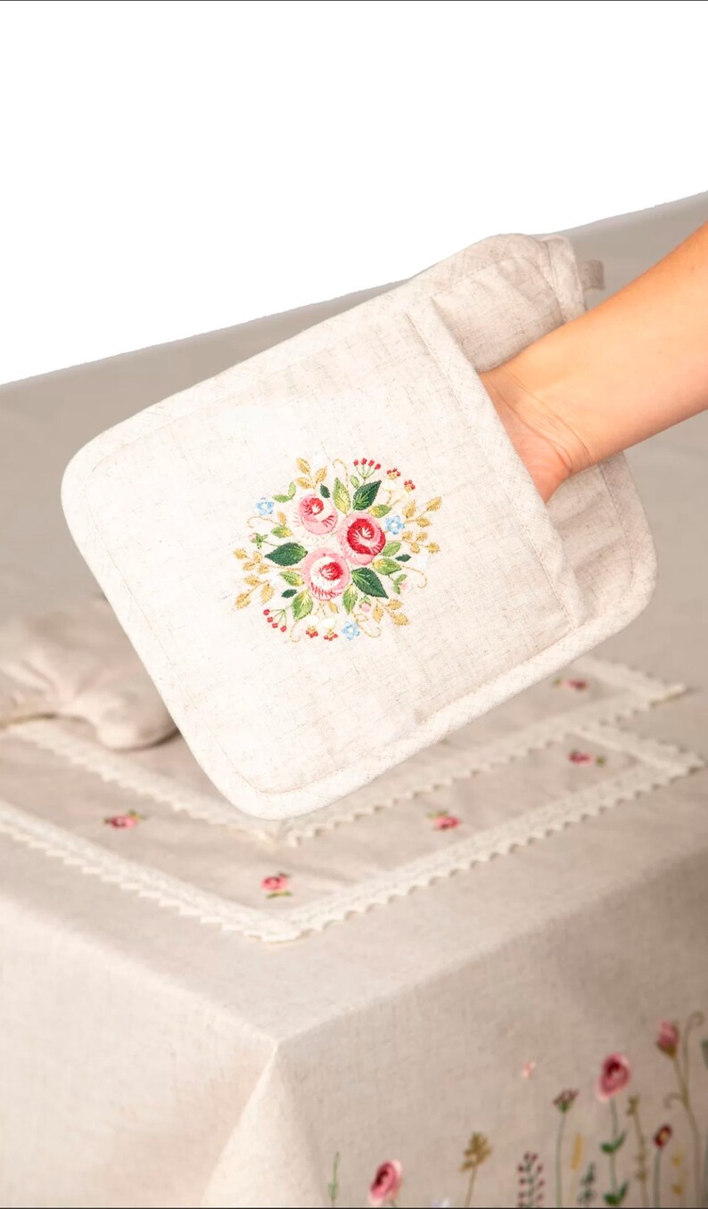 May include: Beige oven mitt with an embroidered floral design featuring pink roses, green leaves, and blue accents. The mitt is held over a matching tablecloth with embroidered flowers and lace trim, creating a cohesive kitchen textile set.