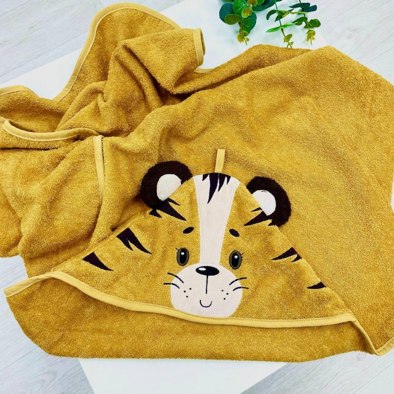 May include: A yellow hooded towel with a tiger face embroidered on the hood. The tiger has black eyes, a black nose, and black stripes.