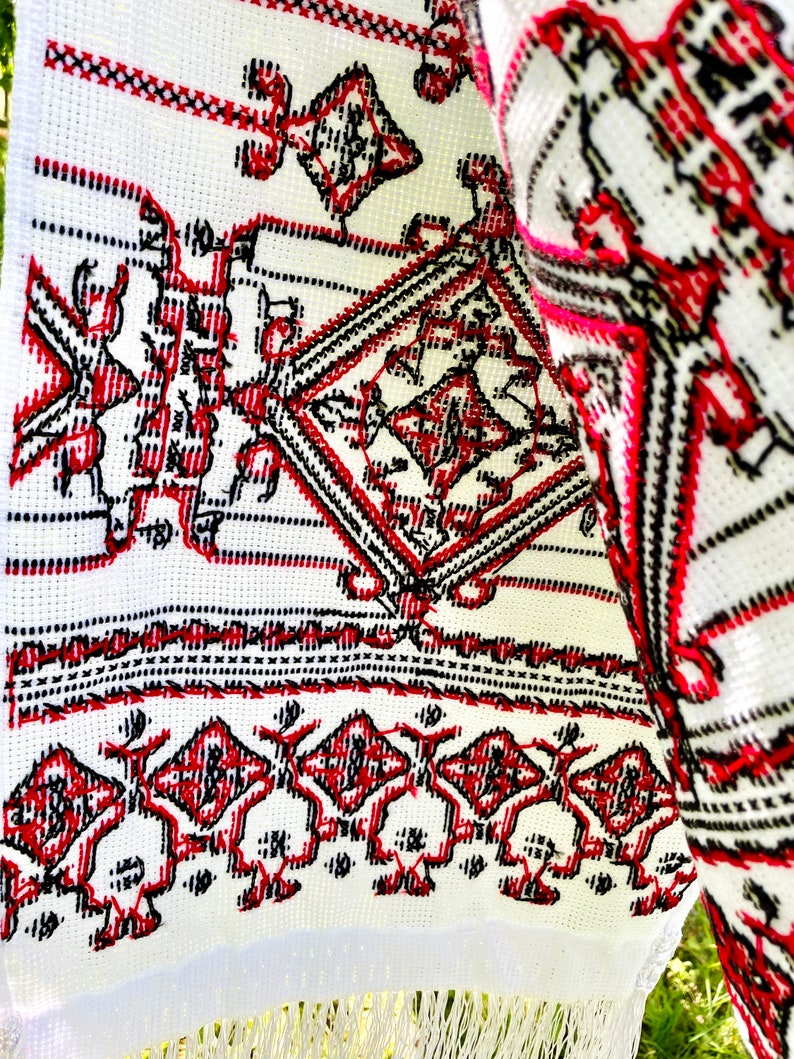 Puede incluir: Textil blanco con intrincados patrones bordados en rojo y negro. El dise&ntilde;o presenta formas geom&eacute;tricas y figuras estilizadas. La tela tiene una franja en el borde inferior. El bordado es detallado y parece hecho a mano.