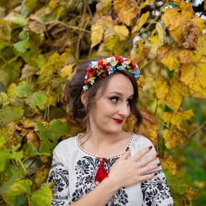 Pode incluir: Uma mulher vestindo uma blusa branca bordada com uma borla vermelha e uma coroa de flores. Ela est&aacute; em p&eacute; em frente a um fundo de folhas amarelas e verdes.