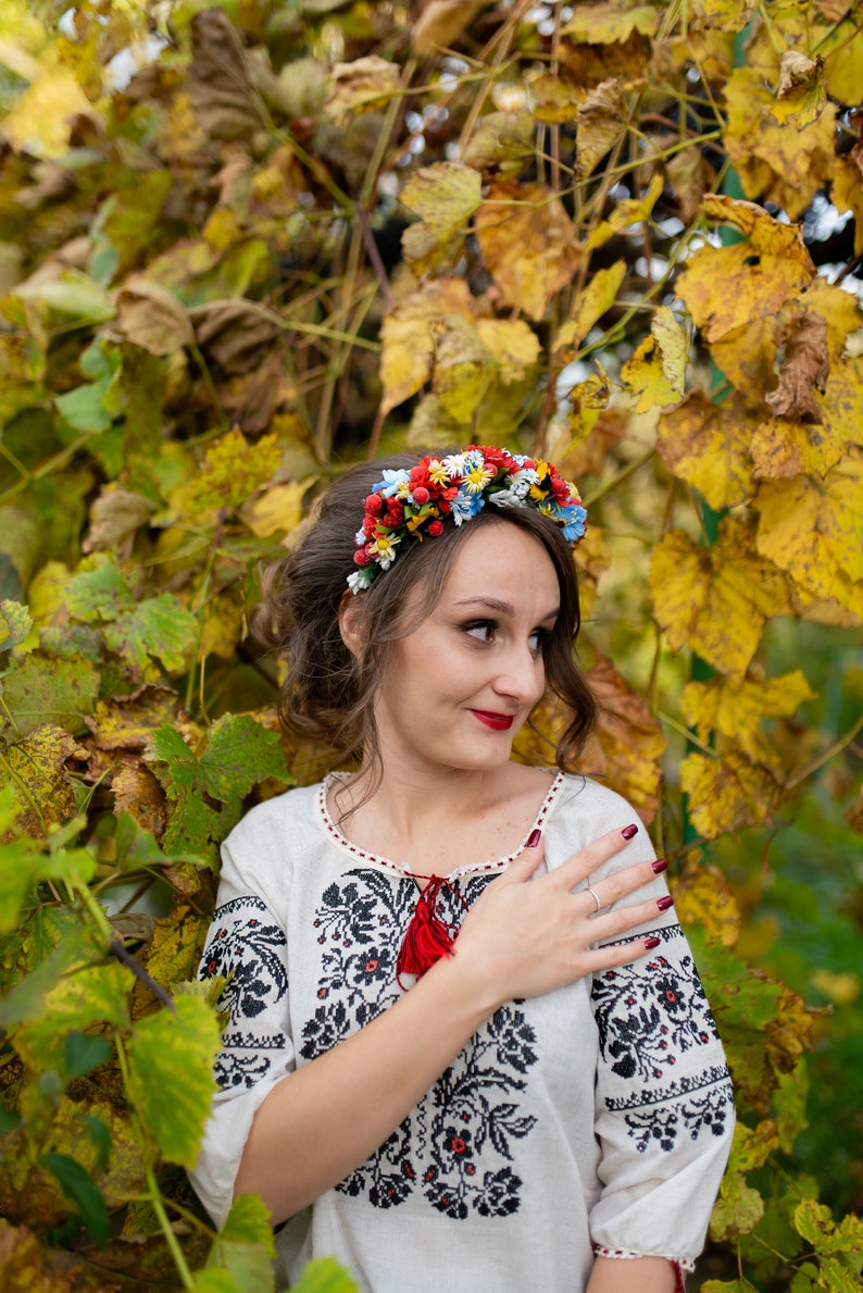 Pode incluir: Uma mulher vestindo uma blusa branca bordada com uma borla vermelha e uma coroa de flores. Ela est&aacute; em p&eacute; em frente a um fundo de folhas amarelas e verdes.
