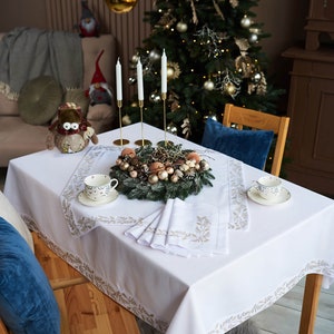 Puede incluir: Una mesa festiva con un mantel blanco con un borde dorado de hojas. La mesa est&aacute; decorada con tazas de t&eacute;, servilletas y una corona decorativa. Candelabros dorados y un &aacute;rbol de Navidad completan la escena.