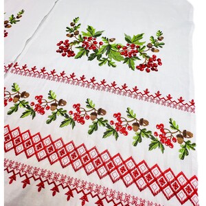K&ouml;nnte beinhalten: Wei&szlig;er Stoff mit gestickten roten Beeren, gr&uuml;nen Bl&auml;ttern und braunen Eicheln. Das Design umfasst geometrische Muster und dekorative Bord&uuml;ren in roter Farbe, wodurch eine Volkskunst-&Auml;sthetik entsteht.