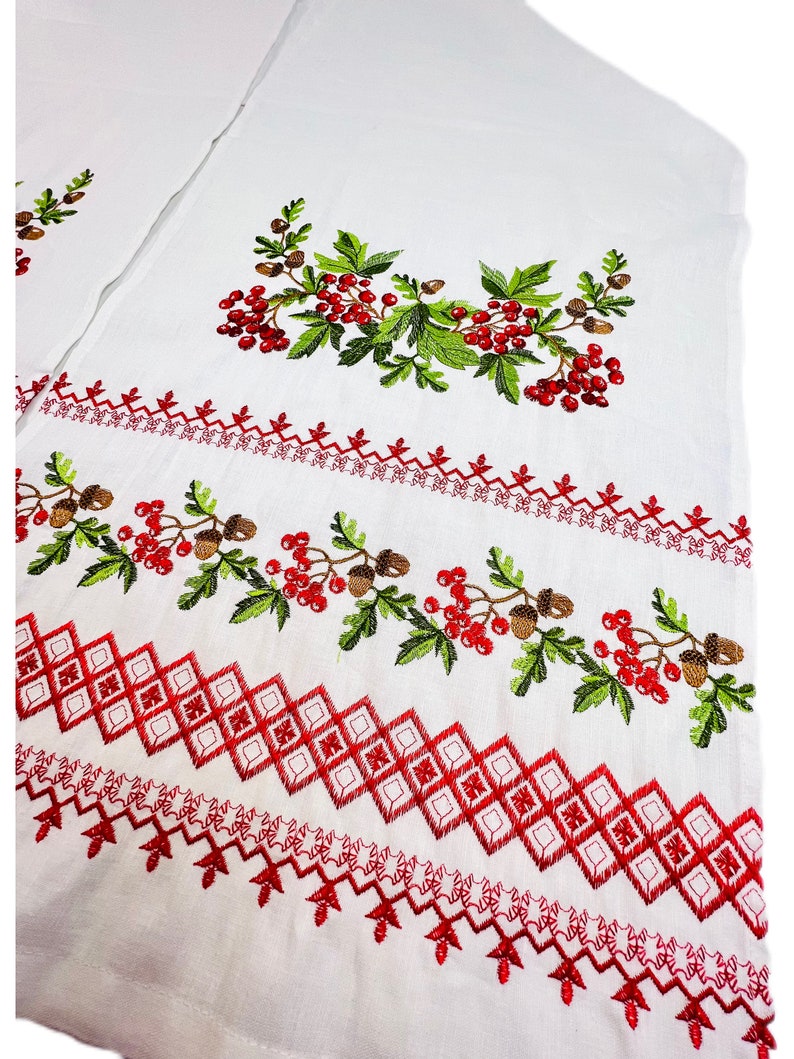 K&ouml;nnte beinhalten: Wei&szlig;er Stoff mit gestickten roten Beeren, gr&uuml;nen Bl&auml;ttern und braunen Eicheln. Das Design umfasst geometrische Muster und dekorative Bord&uuml;ren in roter Farbe, wodurch eine Volkskunst-&Auml;sthetik entsteht.