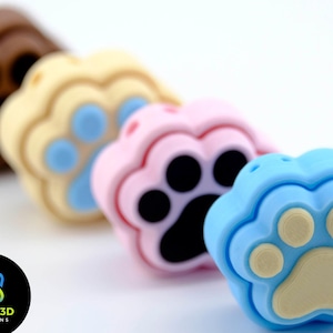 Op de afbeelding: Vier kleurrijke siliconen hangers in de vorm van een poot in bruin, beige, roze en blauw. De hangers zijn in een rij van links naar rechts gerangschikt.