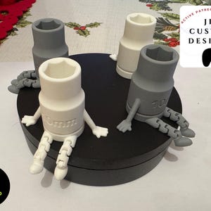 Puede incluir: Cuatro llaves de vaso impresas en 3D con piernas y brazos articulados, sentadas sobre una base circular negra. Las llaves están pintadas en blanco y gris, con los tamaños 10 mm y 6 mm impresos en ellas. La base tiene el texto "Will's 3D Solutions" impreso en ella.