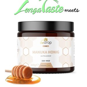 May include: A jar of Manuka honey with a wooden honey dipper. The jar has a black lid and a label with the text "MANUKA HONIG" and "500+ MGO". The honey dipper is resting on a small puddle of honey. The text "Longa Taste meets" is at the top.