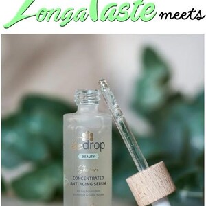 May include: A clear glass bottle of Bedrop Beauty concentrated anti-aging serum with a dropper. The bottle has a light green label with gold text and a wooden cap. The text "Longa Taste meets" is at the top.