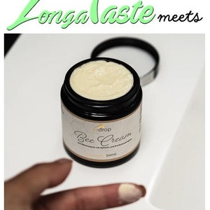 May include: A black jar of Bee Cream with a white label and a light beige cream inside. The jar is open, and the lid is next to it. The text "Longa Taste meets" is above the jar in green and black.