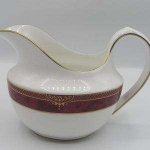 May include: White ceramic creamer with a burgundy band and gold decorative pattern. The creamer has a curved spout and a looped handle. Gold trim accents the rim and handle. A classic piece for serving cream or milk.