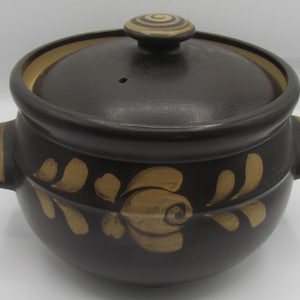 May include: A dark brown ceramic cooking pot with a lid and two handles. The pot is decorated with a floral design in a lighter brown colour. The lid has a small hole and a decorative knob.