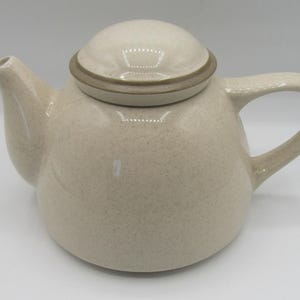 May include: A beige ceramic teapot with a matching lid. The teapot has a rounded body, a curved handle, and a spout. The lid is topped with a brown ring. The teapot has a speckled pattern.