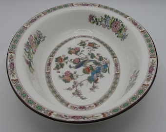 May include: A white ceramic bowl with a brown rim and a decorative floral and bird design. The bowl features a central medallion with a bird and floral motif, surrounded by a patterned border with green and brown accents. The outer rim has additional floral details.