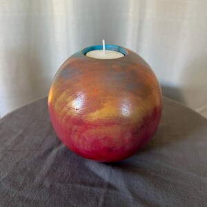 May include: A round, red, gold, and blue candle holder with a white candle inside. The holder is made of wood and has a smooth, polished finish.