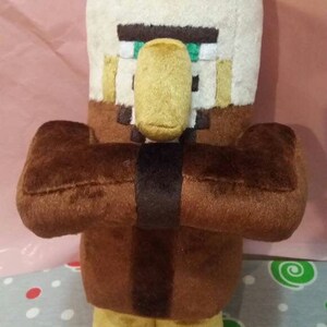 May include: A plush toy of a Minecraft Villager character. The toy has a large head with a beige top and a tan face with a yellow nose. The body is brown with brown arms and yellow feet. A black band wraps around the torso.
