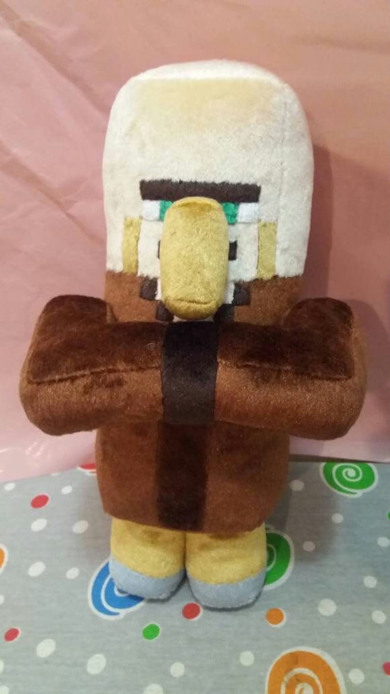 May include: A plush toy of a Minecraft Villager character. The toy has a large head with a beige top and a tan face with a yellow nose. The body is brown with brown arms and yellow feet. A black band wraps around the torso.