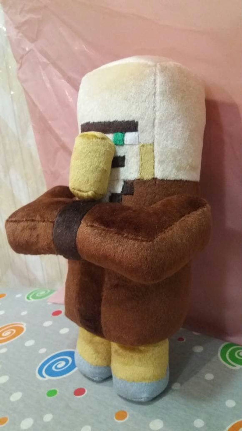 May include: A plush toy of a Minecraft Villager character. The toy is primarily brown, with a white head and a yellow nose. It has a small, cylindrical yellow object held in its arms. The toy is standing on a patterned surface.