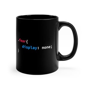 May include: A black ceramic mug featuring white, red, and blue CSS code text that reads .fear{ display: none; }. The mug has a cylindrical body with a curved handle on the right side, presenting a smooth, glossy finish throughout the entire exterior.