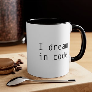 May include: A white ceramic mug with a black interior and a black handle. The exterior features the black text "I dream in code" printed in a simple sans-serif font. The mug is positioned on a light-colored wooden surface next to a silver spoon.