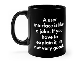 May include: A black ceramic mug with a handle on the left side. The mug features white text that reads: A user interface is like a joke. If you have to explain it, its not very good. The design is printed clearly on the front of the black mug.