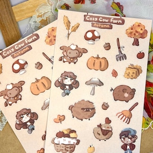 May include: A sheet of stickers featuring a cozy cow farm theme. The stickers are illustrated in a cartoon style and feature a variety of autumnal elements, including cows, pumpkins, mushrooms, a pig, a chicken, a basket of apples, and a farmhand. The stickers are printed on a beige background with the text "Cozy Cow Farm Autumn" at the top.