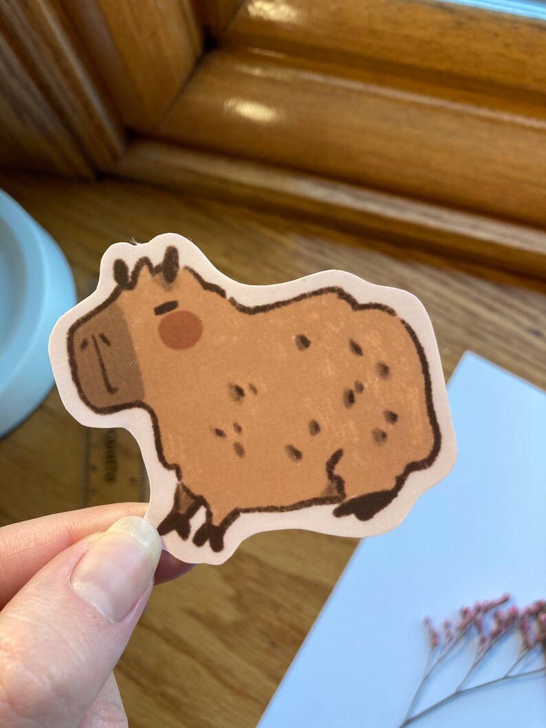 May include: A brown and white capybara sticker with a cartoon-like style. The capybara is facing the left side of the image and has a pink nose.