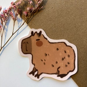 May include: A brown and white cartoon capybara sticker with black eyes and a brown nose. The capybara is sitting on its hind legs with its front paws in the air.