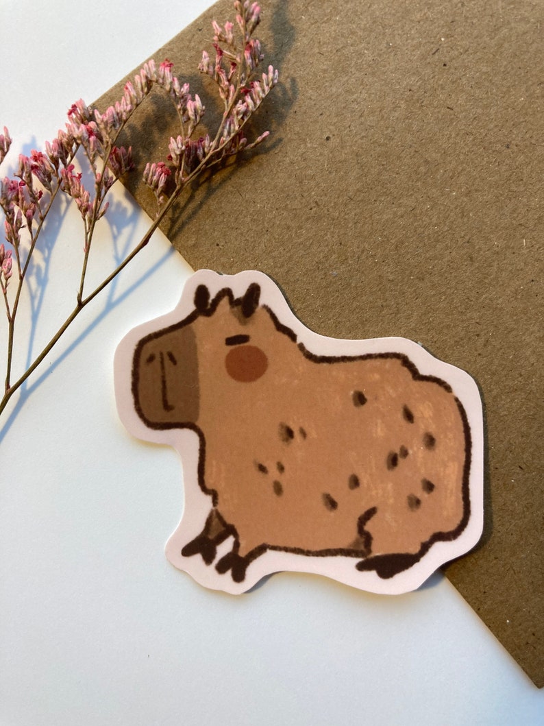 May include: A brown and white cartoon capybara sticker with black eyes and a brown nose. The capybara is sitting on its hind legs with its front paws in the air.