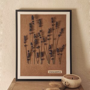 Peut inclure: Une œuvre d'art encadrée avec des tiges de lavande séchées aux fleurs violettes sur un fond marron. L'œuvre est dans un cadre noir et repose sur une surface en bois. Une petite bougie et des plantes séchées sont également sur la surface.