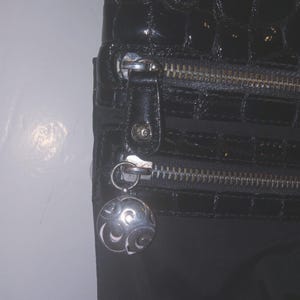 May include: Black handbag with a glossy, textured finish. Features a silver zipper and a decorative silver charm with a swirling design. The bag has a rectangular shape and a dark, neutral color.