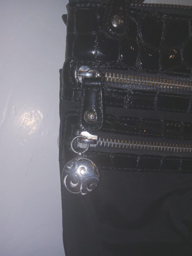 May include: Black handbag with a glossy, textured finish. Features a silver zipper and a decorative silver charm with a swirling design. The bag has a rectangular shape and a dark, neutral color.