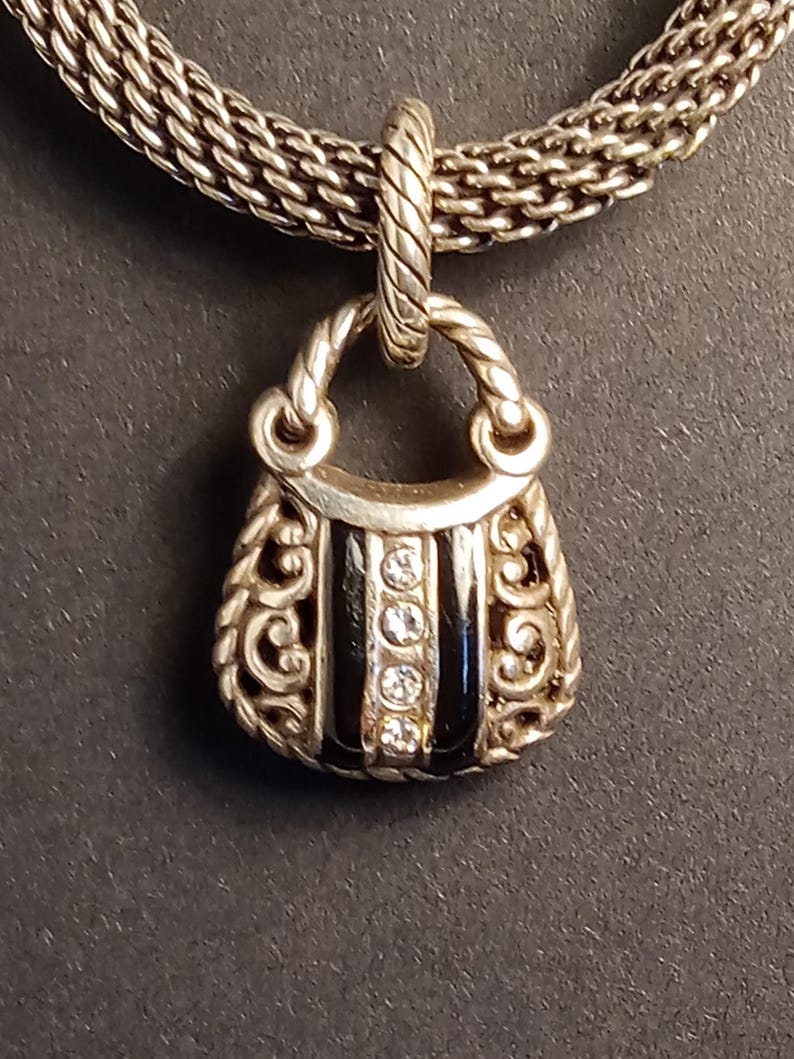 Pu&ograve; includere: Collana con pendente a forma di borsa argentata. Il pendente presenta un piccolo design a forma di borsa con strisce di smalto nero e pietre chiare scintillanti. La catena della collana &egrave; in stile intrecciato.