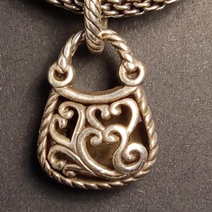 Pu&ograve; includere: Una collana con pendente argentato con un ciondolo a forma di borsa. Il ciondolo ha un design traforato decorativo con un bordo a corda e un piccolo anello per la catena. La catena &egrave; in stile intrecciato.