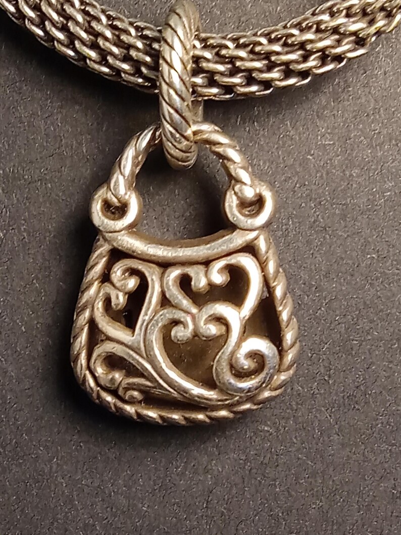 Pu&ograve; includere: Una collana con pendente argentato con un ciondolo a forma di borsa. Il ciondolo ha un design traforato decorativo con un bordo a corda e un piccolo anello per la catena. La catena &egrave; in stile intrecciato.