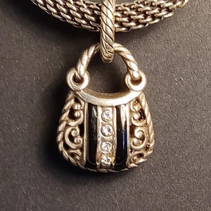 Pu&ograve; includere: Collana con pendente a forma di borsa argentata. Il pendente presenta un piccolo design a forma di borsa con strisce di smalto nero e tre pietre preziose trasparenti. La catena della collana &egrave; in stile intrecciato. Il pendente ha volute decorative.