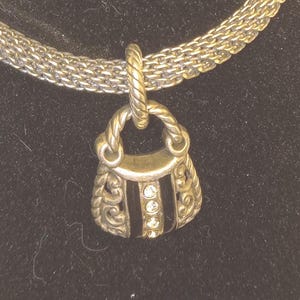 Pu&ograve; includere: Collana color argento con un ciondolo a forma di borsetta. Il ciondolo presenta una striscia nera con tre gemme rotonde e trasparenti. La borsetta ha decorazioni a volute e un manico intrecciato.