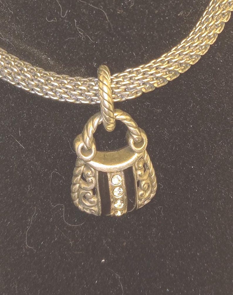 Pu&ograve; includere: Collana color argento con un ciondolo a forma di borsetta. Il ciondolo presenta una striscia nera con tre gemme rotonde e trasparenti. La borsetta ha decorazioni a volute e un manico intrecciato.
