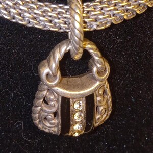 Pu&ograve; includere: Collana color argento con un ciondolo a forma di borsa. Il ciondolo ha una striscia centrale nera con tre gemme trasparenti. La catena &egrave; un mix di maglie argentate e dorate, creando un effetto bicolore. Lo sfondo &egrave; nero.