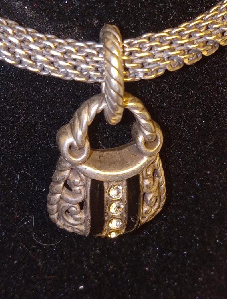 Pu&ograve; includere: Collana color argento con un ciondolo a forma di borsa. Il ciondolo ha una striscia centrale nera con tre gemme trasparenti. La catena &egrave; un mix di maglie argentate e dorate, creando un effetto bicolore. Lo sfondo &egrave; nero.