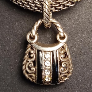 Pu&ograve; includere: Collana color argento con un ciondolo a forma di borsetta. Il ciondolo presenta strisce di smalto nero e tre pietre preziose rotonde e trasparenti. La borsetta ha volute ornate.