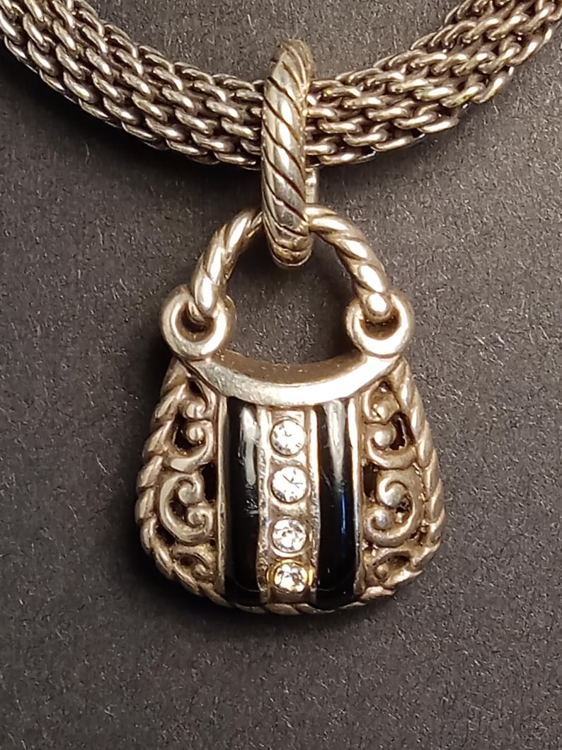 Pu&ograve; includere: Collana color argento con un ciondolo a forma di borsetta. Il ciondolo presenta strisce di smalto nero e tre pietre preziose rotonde e trasparenti. La borsetta ha volute ornate.
