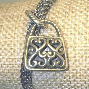 Pu&ograve; includere: Una collana con pendente in metallo argentato con un ciondolo a forma di lucchetto decorativo. Il ciondolo presenta un design traforato ornato da motivi a volute. La catena della collana &egrave; in stile intrecciato e il pendente &egrave; sospeso da un'ansa ritorta.