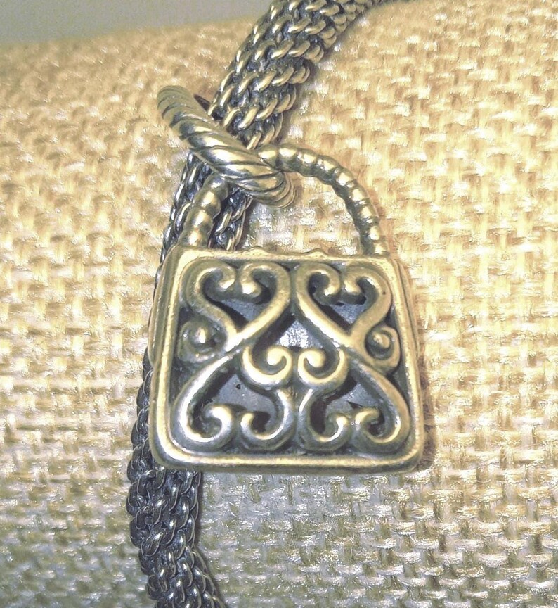 Pu&ograve; includere: Una collana con pendente in metallo argentato con un ciondolo a forma di lucchetto decorativo. Il ciondolo presenta un design traforato ornato da motivi a volute. La catena della collana &egrave; in stile intrecciato e il pendente &egrave; sospeso da un'ansa ritorta.